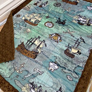 May include: A baby blanket featuring a pirate map design with brown ships, a skull and crossbones, and nautical elements. The blanket has a brown, textured border and is made of soft fabric.