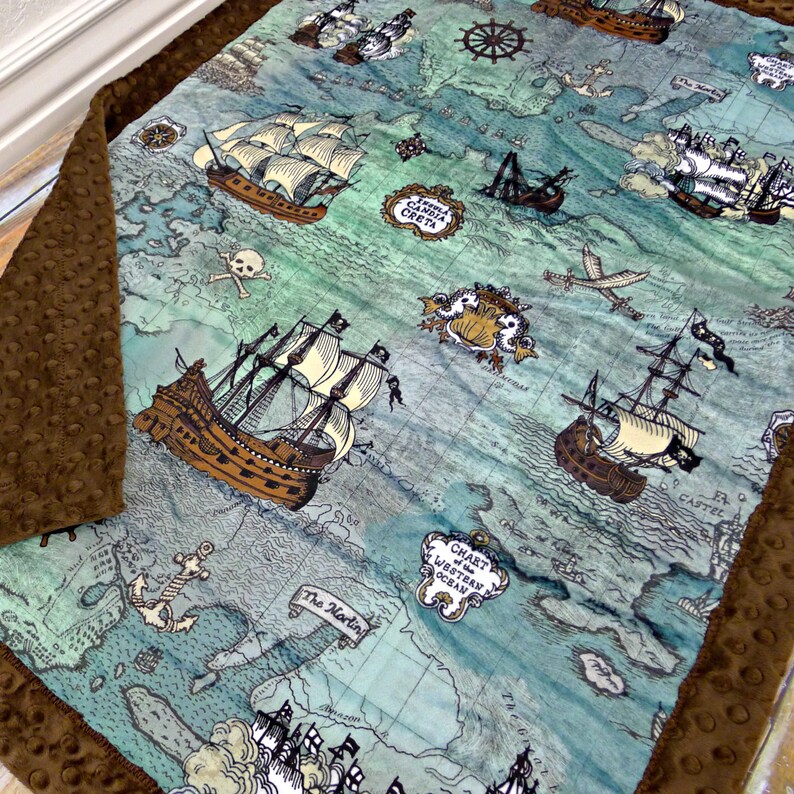 May include: A baby blanket featuring a pirate map design with brown ships, a skull and crossbones, and nautical elements. The blanket has a brown, textured border and is made of soft fabric.