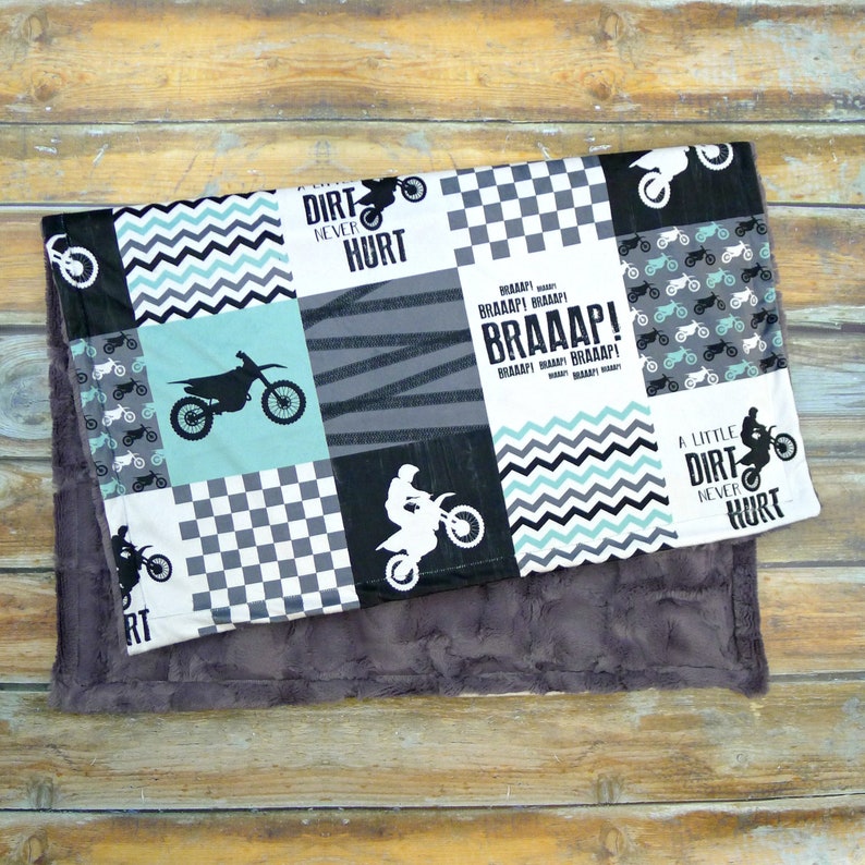 May include: A gray and white baby blanket with a patchwork design featuring motocross bikes, checkered patterns, and the text "Dirt Never Hurt" and "Braaap!" in black.