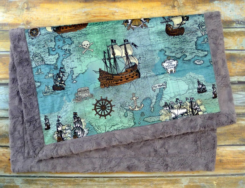 May include: A baby blanket featuring a pirate map design with ships, a skull and crossbones, and the text "Chart of the Western Ocean". The blanket has a soft, gray, plush border.