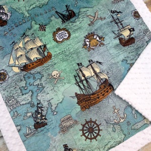 May include: A baby blanket featuring a map design with illustrations of pirate ships, anchors, and a skull and crossbones. The blanket has a white, textured border and a teal and blue color scheme.