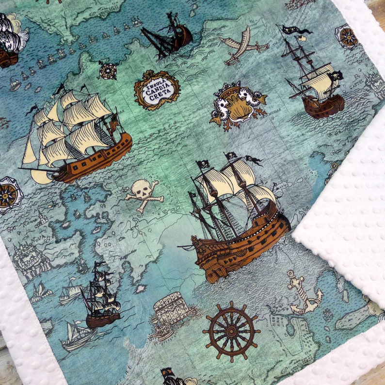 May include: A baby blanket featuring a map design with illustrations of pirate ships, anchors, and a skull and crossbones. The blanket has a white, textured border and a teal and blue color scheme.