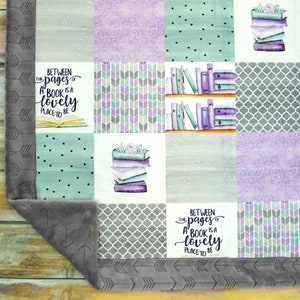 Pode incluir: Uma colcha de retalhos em tons de cinza, branco e roxo. A colcha apresenta uma variedade de padr&otilde;es, incluindo flechas, pontos e um design de treli&ccedil;a. A colcha tamb&eacute;m apresenta uma pilha de livros com flores no topo e o texto "Between the pages of a book is a lovely place to be".