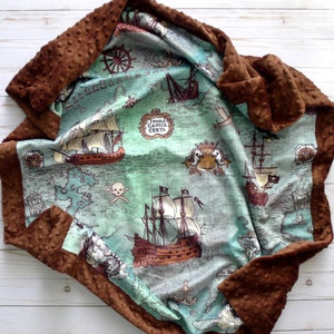 May include: A baby blanket with a pirate map design. The blanket features a teal and brown map with illustrations of pirate ships, compasses, and skulls. The border is a textured brown fabric.