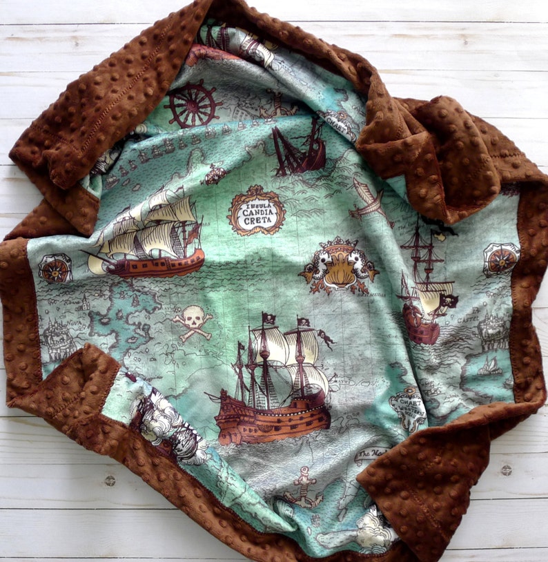 May include: A baby blanket with a pirate map design. The blanket features a teal and brown map with illustrations of pirate ships, compasses, and skulls. The border is a textured brown fabric.