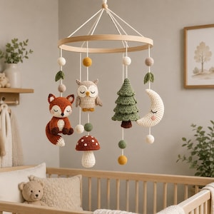 Può includere: Una giostrina sospesa per la cameretta con figure all'uncinetto di una volpe, un gufo, un abete, una luna crescente e un fungo. Gli elementi sono appesi a una cornice circolare in legno tramite cordini bianchi e decorati con piccole sfere.