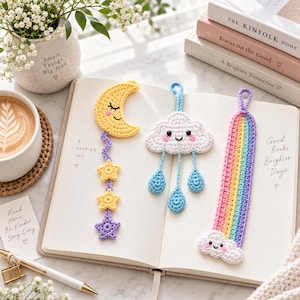 May include: Three crocheted bookmarks resting on an open book. One is a yellow crescent moon with three dangling stars. The second is a white cloud with three blue raindrops. The third is a vertical rainbow strip ending in a small white cloud.