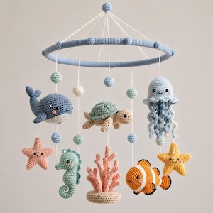 Op de afbeelding: Een gehaakte mobiel voor de babykamer met een blauw cirkelvormig frame en hangende zeedieren, waaronder een blauwe walvis, een groene schildpad, een blauwe kwal, een zeepaardje, een clownvis, twee zeesterren en een roze koraalstuk.