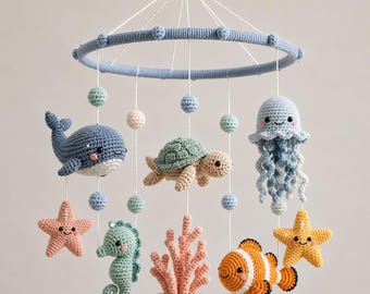 Pode incluir: Um móbile de crochê para quarto de bebê com uma estrutura circular azul e criaturas marinhas penduradas, incluindo uma baleia azul, uma tartaruga verde, uma água-viva azul, um cavalo-marinho, um peixe-palhaço, duas estrelas-do-mar e um coral rosa.