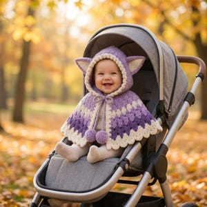 May include: A crochet hooded cape for an infant, featuring a light purple and dark purple gradient pattern with cream-colored trim. The hood includes pointed ears, and the front is secured with a drawstring ending in two matching pom-poms.