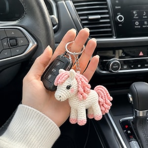 May include: A crochet unicorn keychain featuring a cream-colored body with a pink mane, tail, and hooves. The small stuffed unicorn is attached to a silver-toned metal split ring and chain, positioned next to a black plastic car key fob.