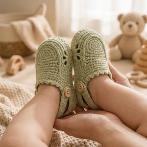 May include: A pair of sage green crocheted baby shoes featuring a textured knit pattern, a scalloped edge along the opening, and a small wooden button on the side strap. The shoes have a rounded toe design and a flexible, soft construction.