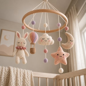 Puede incluir: Un móvil de cuna con un marco circular de madera y figuras de ganchillo colgantes que incluyen un conejo color crema, un globo aerostático multicolor, una nube sonriente, una estrella color melocotón y una luna creciente, con pequeñas esferas.