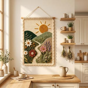 May include: A rectangular crochet wall hanging featuring a landscape scene with a yellow sun, rolling green hills, a winding path, and various colourful flowers including white daisies, a red bloom, and a pink flower, suspended from a wooden rod.