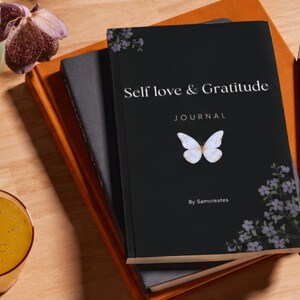 May include: A black journal titled "Self love & Gratitude" with a butterfly illustration and floral accents, resting on a stack of notebooks. A pencil and a glass of orange liquid are also visible on a wooden surface.