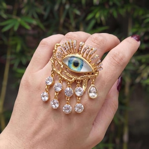 Puede incluir: Un anillo dorado con un motivo de ojo central con iris y pupila azules. El ojo está enmarcado por rayos dorados incrustados con pequeños cristales transparentes. Debajo cuelgan varias cadenas doradas, cada una con un cristal redondo.