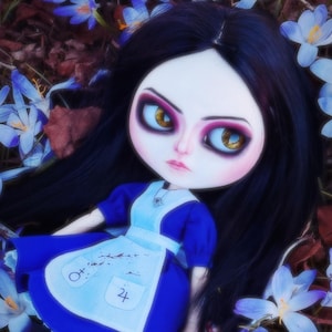 May include: A doll with black hair and large eyes, dressed in a blue dress and white apron, lies among blue and white flowers. The doll's expression is serious, and the overall aesthetic is whimsical and slightly gothic.