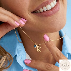 May include: A gold-toned chain necklace featuring a small, faceted blue heart-shaped charm and a gold-toned letter K pendant. The necklace is worn around the neck, showcasing the delicate metal chain and the two distinct hanging charms.