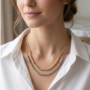 Op de afbeelding: Twee gelaagde kettingen. De bovenste ketting is goudkleurig en de onderste ketting is zilverkleurig. Beide kettingen hebben een reeks cilindrische en bolvormige kralen. De kettingen worden gedragen over een wit overhemd met knopen.