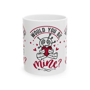 May include: White ceramic mug featuring a cartoon voodoo doll holding a red heart. The mug has the text "Would you be mine?" in a playful font, with red hearts and accents. A fun Valentine's Day gift.