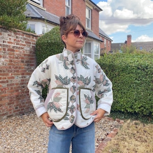 May include: A quilted jacket featuring a cream-coloured base with a repeating pattern of green, pink, and brown fabric leaf appliques. The jacket has a high collar, long sleeves, two large front pockets with olive green trim, and a button-up front.