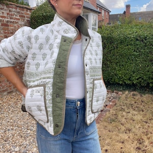 May include: A quilted jacket in white with a green floral block print pattern, featuring a high collar, snap button closure, and two front pockets. The jacket has a solid olive green trim along the placket and cuffs, with a small rectangular label on the lower left pocket that reads Velvet Club.