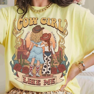 May include: A pale yellow t-shirt featuring a graphic print of two figures in cowboy hats standing in a desert landscape with cacti and roses. The text reads COW GIRL and I KNEW YOU I KNEW YOU and LIKE ME. The design includes ornate gold flourishes.