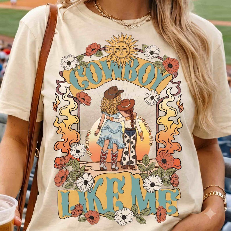 May include: A cream-colored t-shirt featuring a graphic print of two figures in cowboy hats standing back-to-back. The design includes a sun, flowers, and the text "COWBOY" at the top and "I KNEW IT" and "LIKE ME" in blue and yellow lettering.