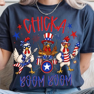 Puede incluir: Camiseta azul marino con un dise&ntilde;o de tres pollos tocando instrumentos musicales y fuegos artificiales. Los textos "CHICKA" y "BOOM BOOM" son en rojo y azul. Los pollos visten atuendos patri&oacute;ticos.
