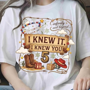 May include: A white t-shirt featuring a graphic print with a brown scroll banner that reads I KNEW IT, I KNEW YOU, the number 5, and the text Cinfinity and beyond. The design includes cowboy boots, a red hat, alphabet blocks, and sheriff stars.