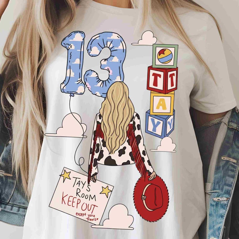 May include: A white t-shirt featuring a graphic of a person with long blonde hair, wearing a cow-print jacket with red fringe. The design includes a blue cloud-patterned number 13 balloon, stacked alphabet blocks spelling TAY, and a red cowboy hat.