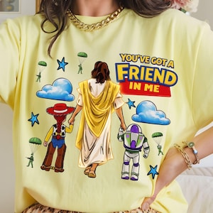 May include: A light yellow t-shirt featuring a graphic design with the text "YOU'VE GOT A FRIEND IN ME". The design includes illustrations of a figure in a yellow robe, a cowgirl, and a space ranger, with blue clouds, stars, and parachuters.