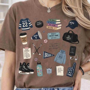 Puede incluir: Camiseta marr&oacute;n con varias ilustraciones, incluyendo una camiseta de hockey, libros, un disco de hockey, una taza de caf&eacute; y una botella de agua. La camiseta tiene el texto "BRIAR U" y "OFF CAMPUS SERIES".