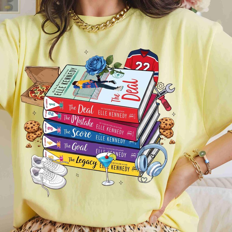 May include: A yellow t-shirt with a design featuring a stack of books titled "The Deal", "The Mistake", "The Score", "The Goal", and "The Legacy" by Elle Kennedy. The design also includes a hockey jersey, pizza, cookies, shoes, and a cocktail.