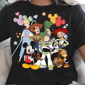 May include: A black short-sleeved t-shirt featuring a colorful graphic print of Mickey Mouse, Woody, Buzz Lightyear, Jessie, Rex, Slinky Dog, and Bo Peep. The design includes various pastel-colored Mickey-shaped balloons and small star icons.