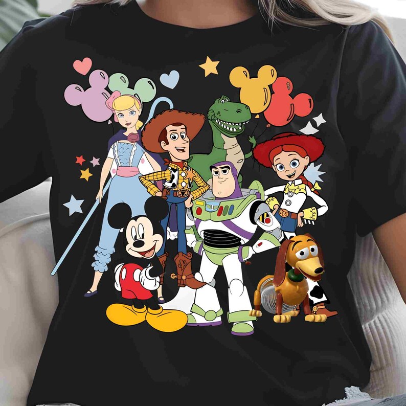 May include: A black short-sleeved t-shirt featuring a colorful graphic print of Mickey Mouse, Woody, Buzz Lightyear, Jessie, Rex, Slinky Dog, and Bo Peep. The design includes various pastel-colored Mickey-shaped balloons and small star icons.