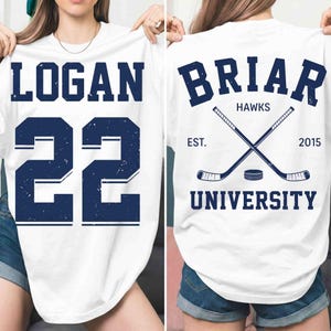 以下が含まれることがあります： 白地にネイビーブルーの文字と数字が入ったTシャツ。「LOGAN」と数字の「22」が前面にプリントされています。背面には「BRIAR HAWKS UNIVERSITY」とクロスしたホッケーのスティックとパック、そして「EST. 2015」と書かれています。
