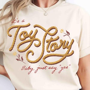 May include: A cream-colored t-shirt featuring the text 'it's a Toy Story' written in a yellow rope-style font, accompanied by the red text 'Baby, just say "yes"' and small floral illustrations in red and yellow on the front of the garment.