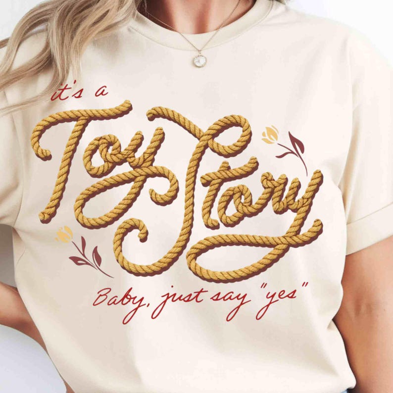 May include: A cream-colored t-shirt featuring the text 'it's a Toy Story' written in a yellow rope-style font, accompanied by the red text 'Baby, just say "yes"' and small floral illustrations in red and yellow on the front of the garment.