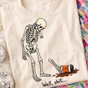 May include: A cream-colored t-shirt featuring a graphic of a skeleton looking down at its detached arm holding a spilled orange cup labeled Pumpkin Spice Coffee, with the text Well, shit... printed in black below the illustration.