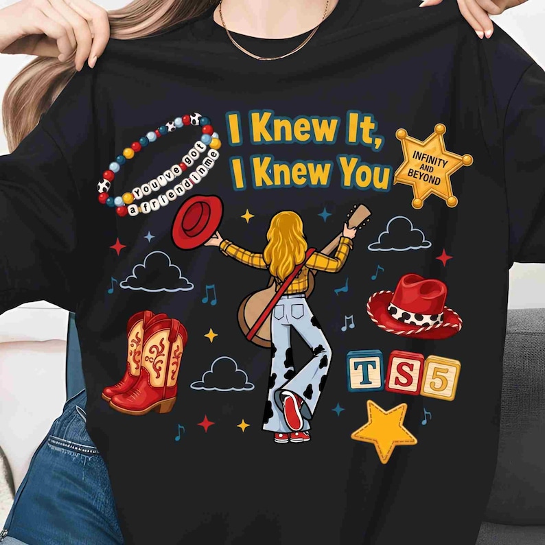May include: A black t-shirt featuring a graphic of a person with blonde hair playing a guitar, wearing a yellow plaid shirt and cow-print pants. The design includes red cowboy boots, a red hat, a sheriff badge, alphabet blocks reading TS5, and text.