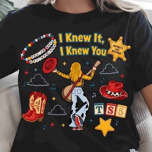 May include: A black t-shirt featuring a graphic of a person with blonde hair holding a guitar, wearing a yellow plaid shirt and cow-print pants. The design includes red cowboy boots, a sheriff badge with text, colorful beads, and the text I Knew It, I Knew You.