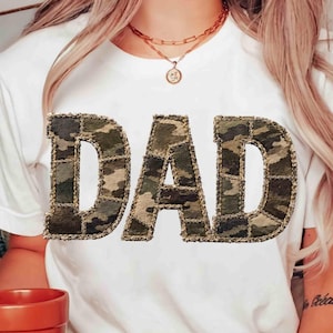 May include: White t-shirt with the word "DAD" in a camouflage pattern. The letters are outlined with a brown stitching detail. The shirt is a casual style.