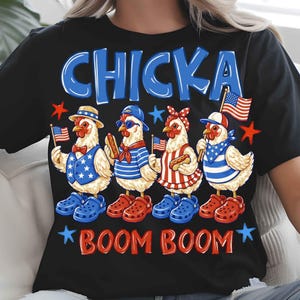 Puede incluir: Camiseta negra con un dise&ntilde;o de cuatro pollos vestidos con atuendos patri&oacute;ticos, sosteniendo banderas americanas y perritos calientes. El texto "CHICKA BOOM BOOM" est&aacute; en azul y rojo.