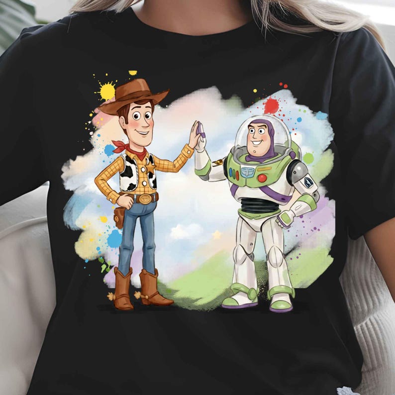May include: A black short-sleeved t-shirt featuring a colorful graphic print of the characters Woody and Buzz Lightyear high-fiving. The design is set against a watercolor-style splash of blue, green, yellow, and red paint splatters.