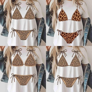 May include: White t-shirts with a leopard print bikini design. The bikini top and bottom are printed in a brown and tan animal print. The bikini top has bow tie straps, and the bottom has tie sides.
