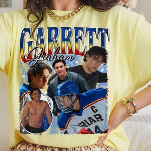Puede incluir: Una camiseta de color amarillo p&aacute;lido con un estampado gr&aacute;fico que incluye el texto GARRETT Graham en una fuente azul y roja. El dise&ntilde;o muestra tres im&aacute;genes de un hombre, una de ellas sin camisa y otra con una camiseta de hockey azul y blanca.