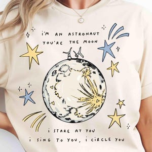 May include: Cream-colored t-shirt with a whimsical illustration featuring a moon, stars, and the text "I'm an astronaut, you're the moon." The design includes a detailed moon graphic and stylized stars in yellow and blue.