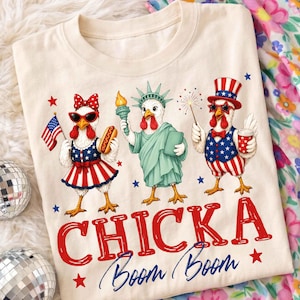 Puede incluir: Camiseta color crema con tres pollos de dibujos animados vestidos con atuendos patri&oacute;ticos. Un pollo sostiene una bandera estadounidense y un perrito caliente, otro est&aacute; vestido como la Estatua de la Libertad, y el tercero lleva un sombrero de copa y una pajarita. El texto "CHICKA Boom Boom" est&aacute; impreso en rojo y azul.