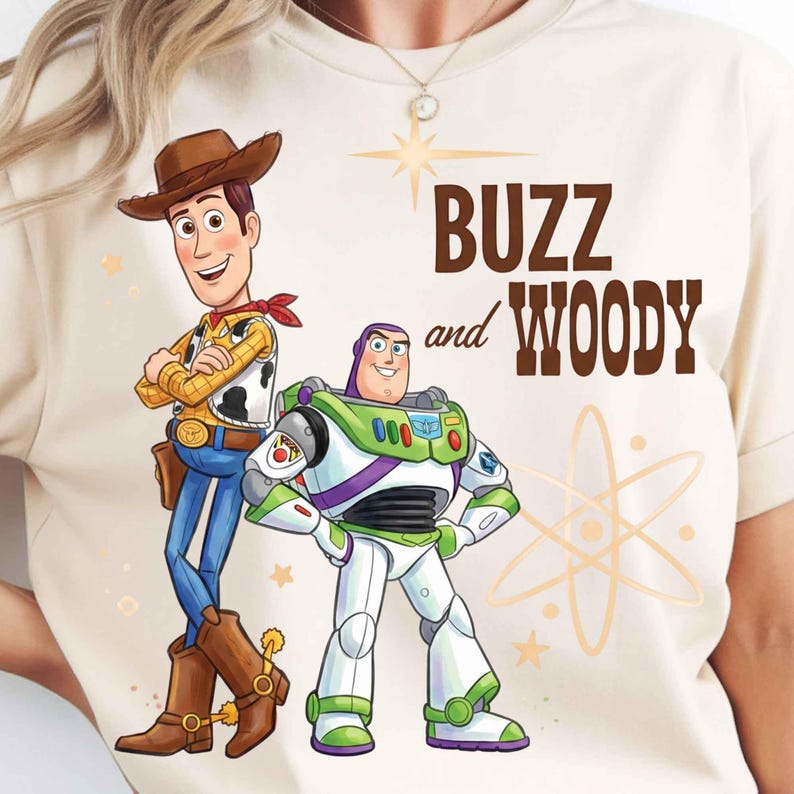 May include: A cream-colored t-shirt featuring a graphic of Woody and Buzz Lightyear. Woody wears a brown cowboy hat, yellow shirt, and blue jeans. Buzz wears a white, green, and purple space suit. The text reads BUZZ and WOODY.